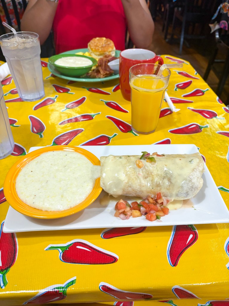 A plate featuring a burrito topped with cheese sauce and diced tomatoes, accompanied by a bowl of grits and a glass of orange juice, set on a vibrant chili pepper-themed tablecloth.