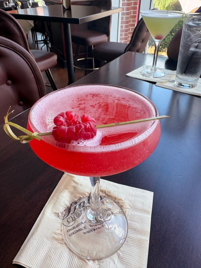 A vibrant red cocktail garnished with a raspberry and served in a stylish glass, placed on a branded napkin. In the background, another drink can be seen.