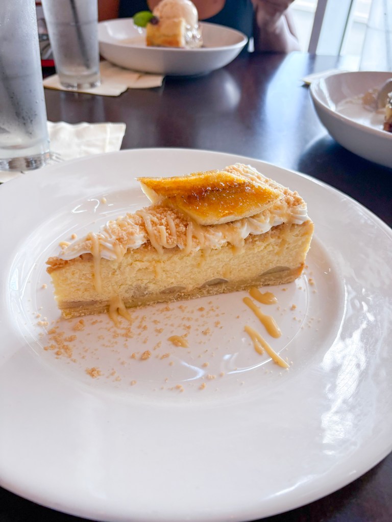 A slice of cheesecake topped with caramelized banana and coconut flakes, served on a white plate with a drizzle of sauce.