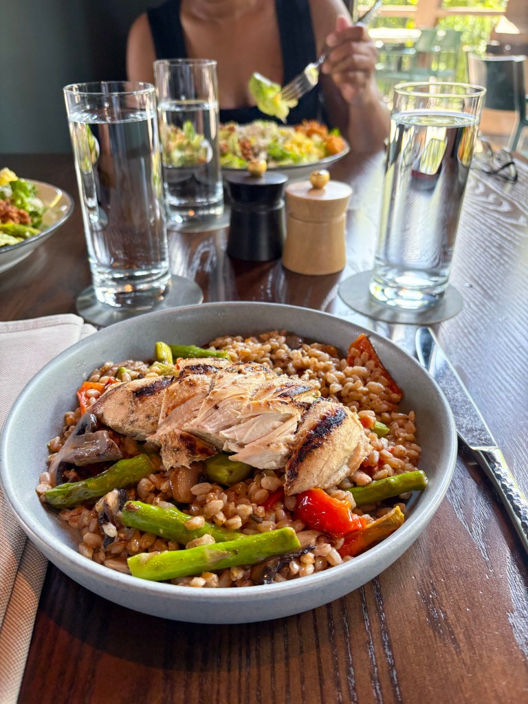 A close-up view of a plate featuring grilled chicken on a bed of farro with asparagus and colorful peppers, accompanied by two glasses of water and a salad in the background.