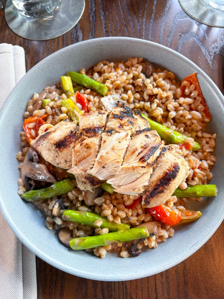 A plate of grilled chicken breast sliced atop a bed of farro, surrounded by colorful vegetables including asparagus, red peppers, and mushrooms.
