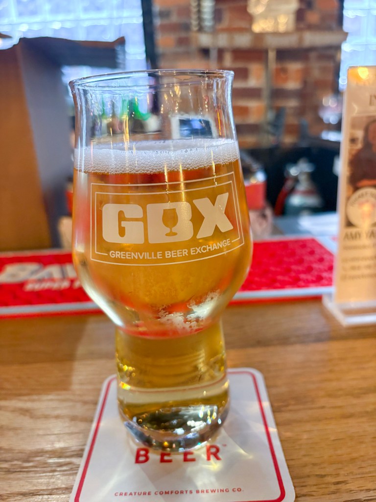 A close-up of a glass of beer with the logo 'GBX' for Greenville Beer Exchange, placed on a coasters with the word 'BEER' visible, set against a blurred bar background.