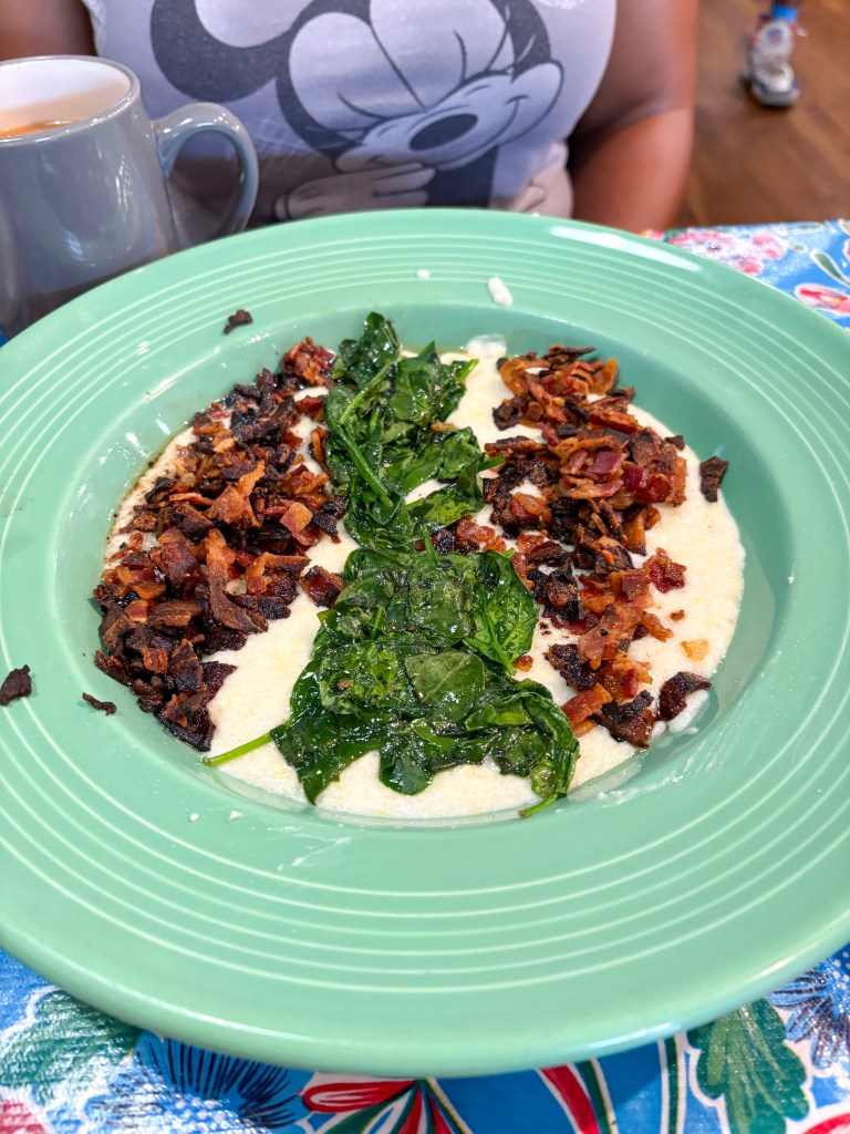 A green plate featuring creamy grits topped with sautéed spinach and crispy bacon bits, with a coffee cup in the background.