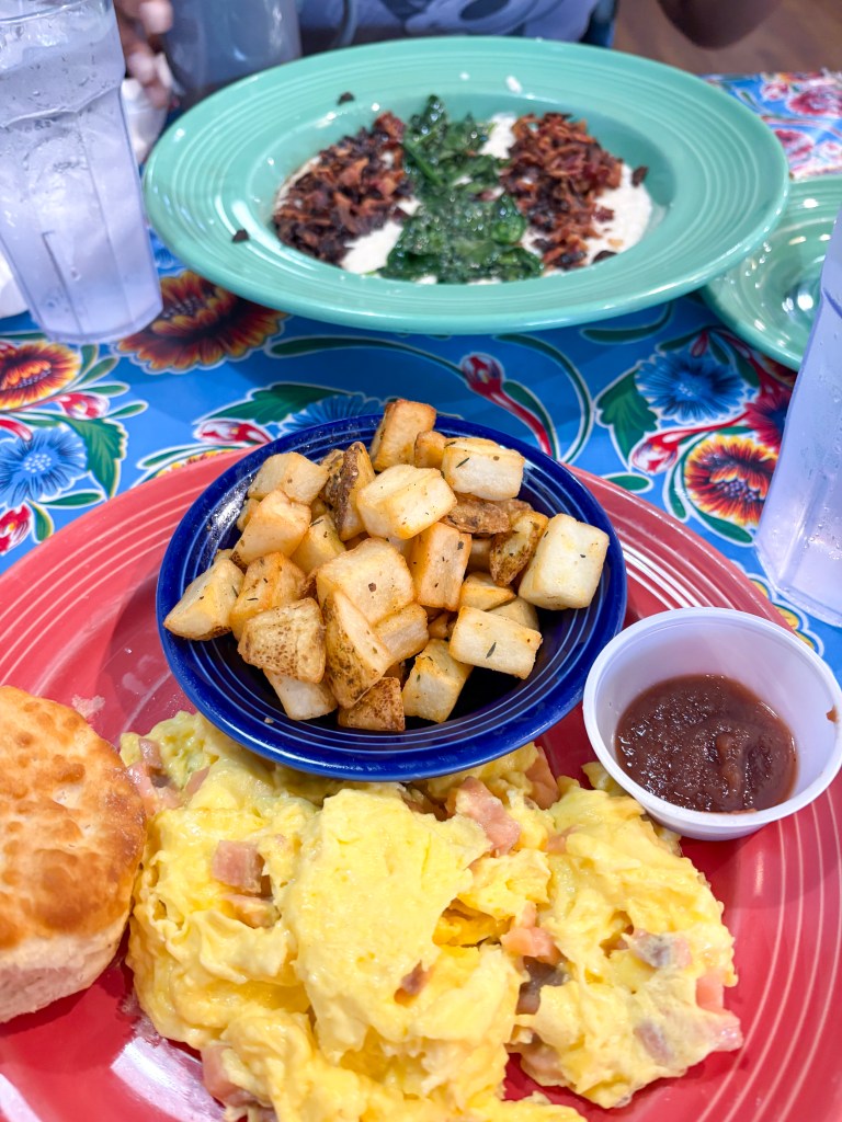 A colorful plate of breakfast featuring scrambled eggs with diced ham, a biscuit, crispy breakfast potatoes in a blue bowl, and a small cup of salsa.